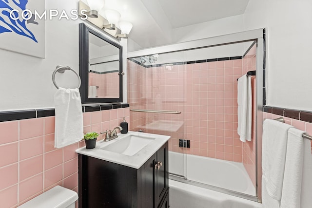 full bathroom featuring tile walls, combined bath / shower with glass door, vanity, toilet, and a wainscoted wall