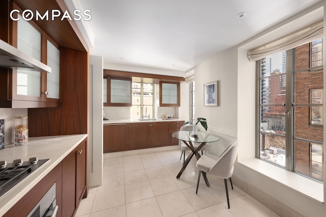 kitchen featuring cooktop, light countertops, glass insert cabinets, a sink, and wall chimney range hood