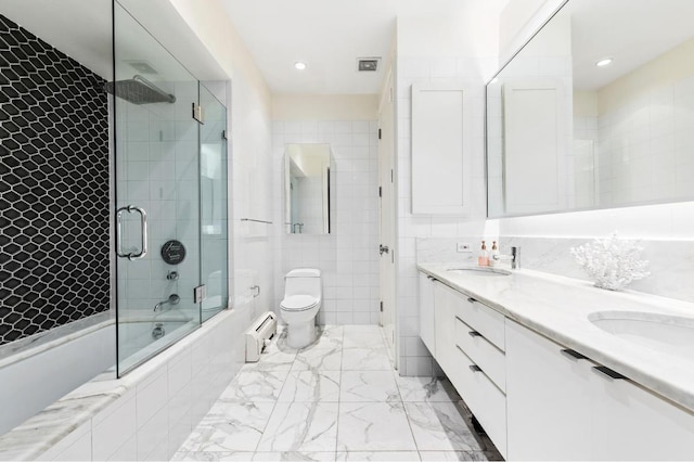 full bathroom with tile walls, a baseboard heating unit, bath / shower combo with glass door, vanity, and toilet