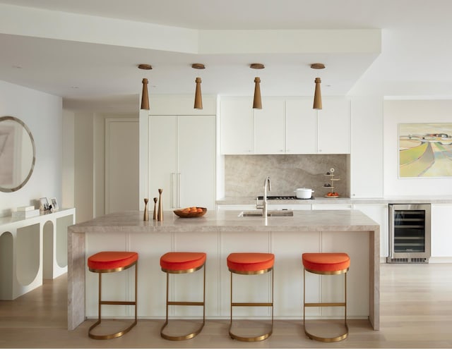 kitchen featuring tasteful backsplash, beverage cooler, white cabinets, light wood-style flooring, and a sink