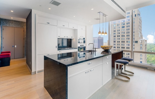 kitchen with sink, hanging light fixtures, light hardwood / wood-style floors, an island with sink, and white cabinets