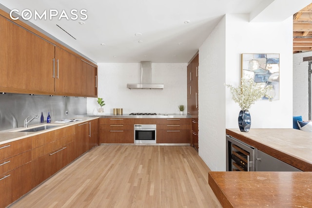 kitchen with brown cabinetry, a sink, oven, wine cooler, and wall chimney exhaust hood