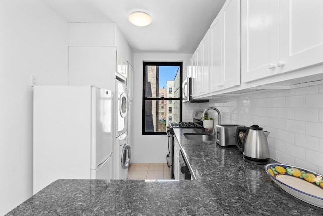 kitchen featuring stainless steel appliances, light tile patterned floors, dark stone counters, stacked washer / dryer, and white cabinets