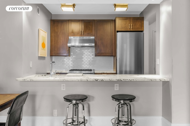 kitchen with range, decorative backsplash, light stone counters, freestanding refrigerator, and under cabinet range hood