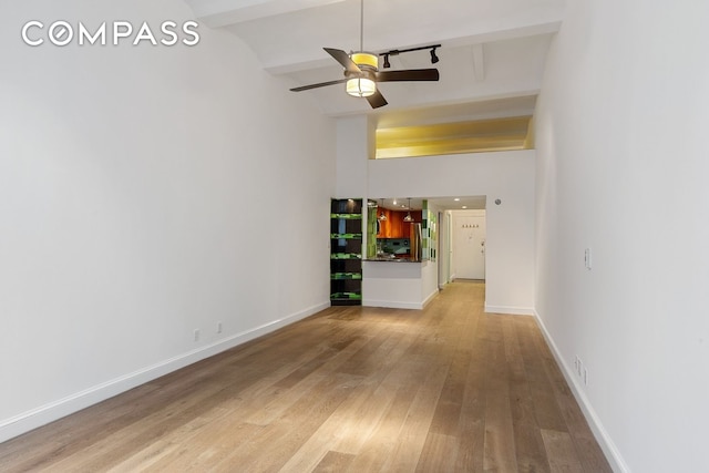 unfurnished living room with beamed ceiling, light wood-style floors, ceiling fan, baseboards, and a high ceiling