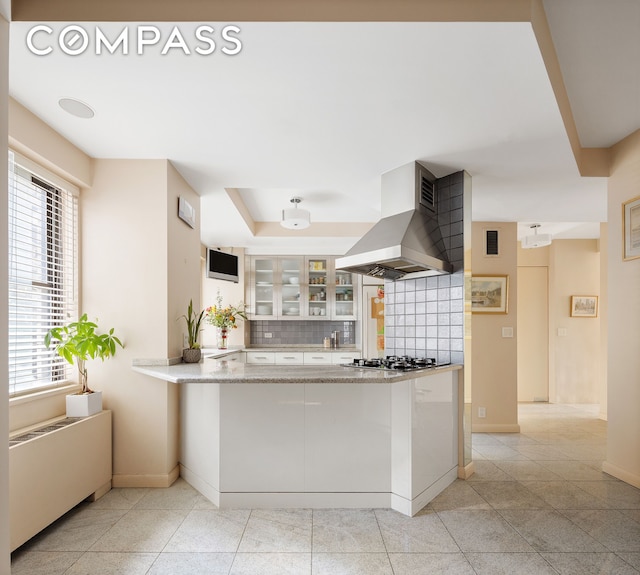 kitchen featuring baseboards, stainless steel gas stovetop, glass insert cabinets, wall chimney exhaust hood, and tasteful backsplash