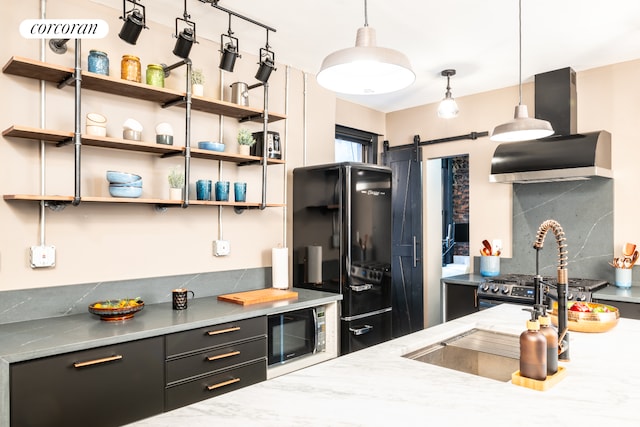 kitchen featuring backsplash, freestanding refrigerator, a barn door, wall chimney range hood, and dark cabinets