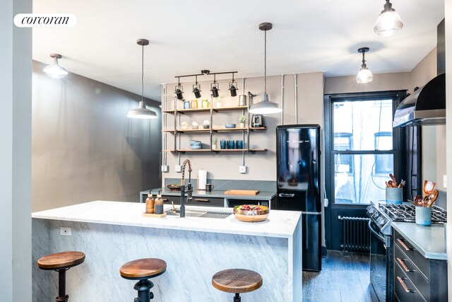 kitchen with stainless steel range with gas cooktop, a kitchen bar, radiator heating unit, freestanding refrigerator, and wall chimney exhaust hood