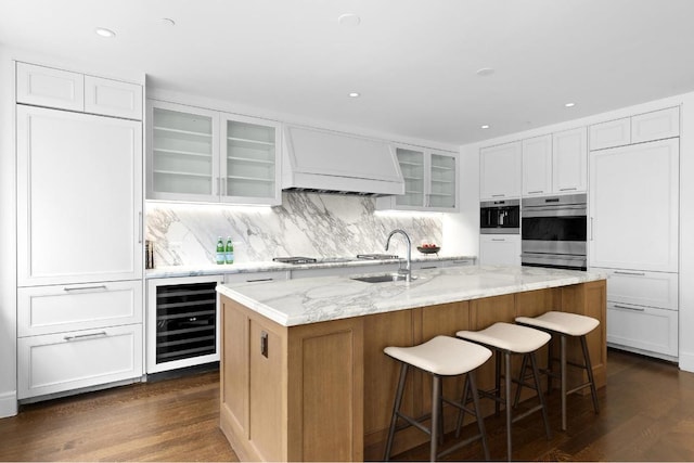 kitchen featuring sink, premium range hood, white cabinetry, a center island with sink, and beverage cooler