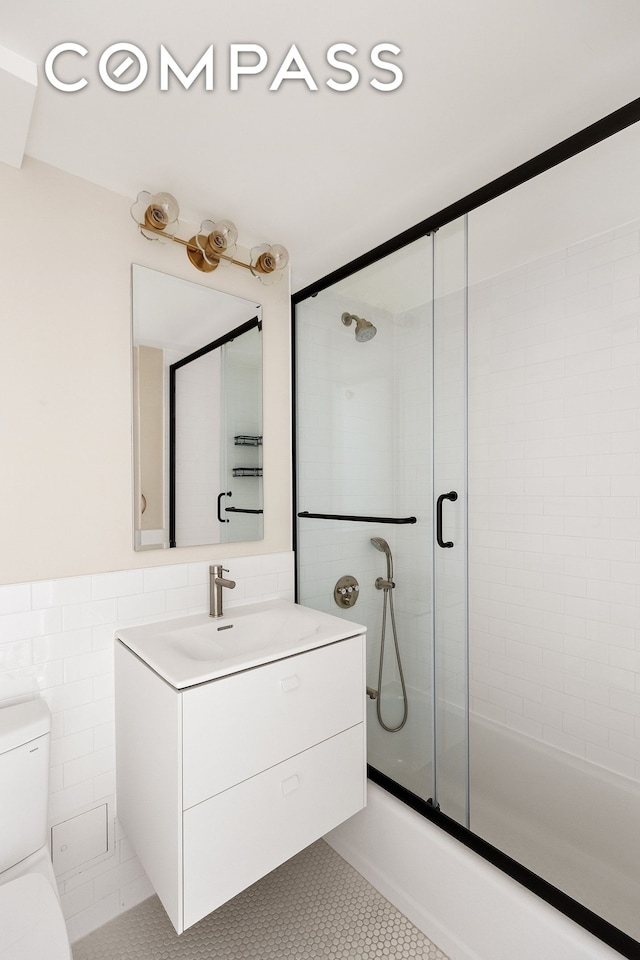 bathroom featuring toilet, tile patterned floors, combined bath / shower with glass door, vanity, and tile walls