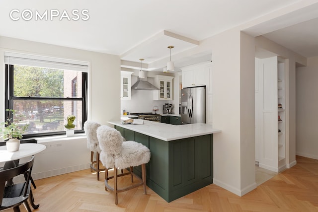 kitchen with a peninsula, white cabinets, wall chimney range hood, stainless steel fridge with ice dispenser, and glass insert cabinets