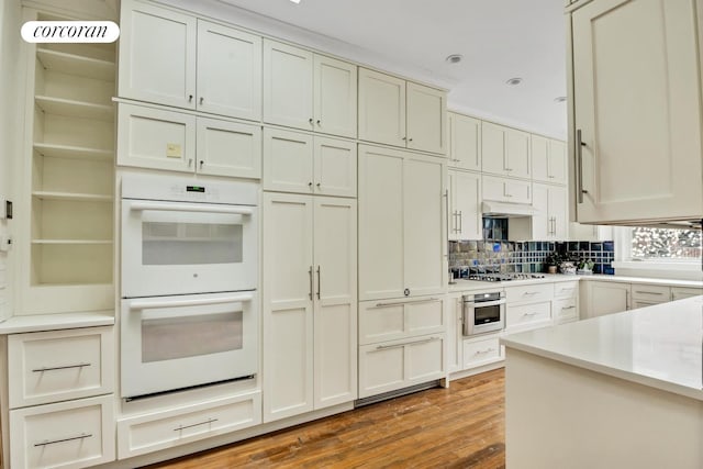 kitchen featuring tasteful backsplash, wood finished floors, light countertops, under cabinet range hood, and double oven