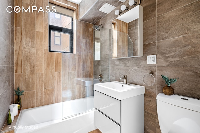 bathroom featuring bath / shower combo with glass door, tile walls, vanity, and toilet