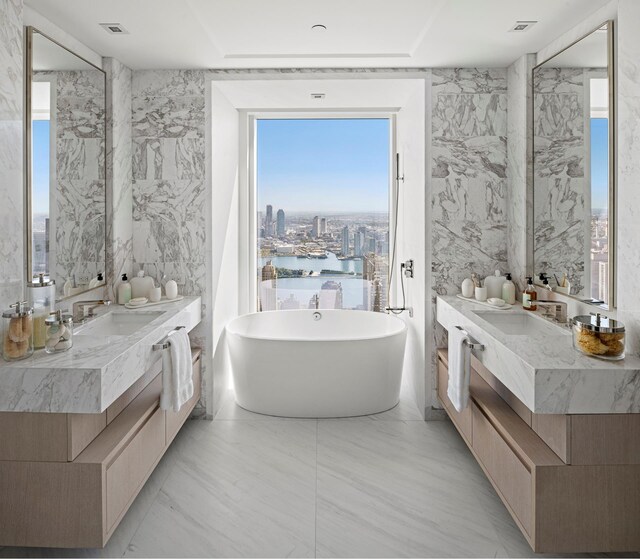 bathroom featuring a freestanding bath, two vanities, a water view, and stone wall