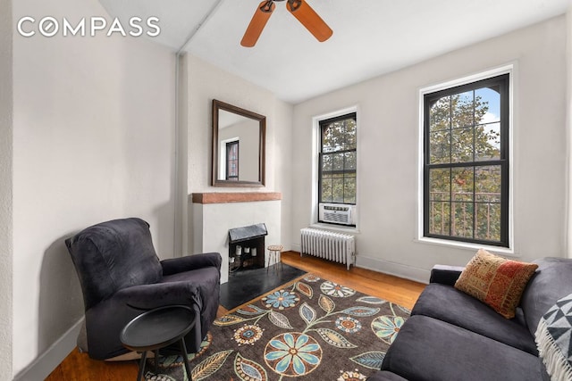 living area with radiator heating unit, cooling unit, ceiling fan, a tiled fireplace, and hardwood / wood-style floors