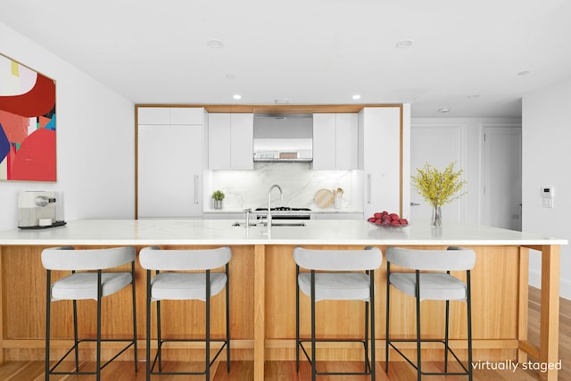 kitchen with white cabinetry, light countertops, wall chimney range hood, decorative backsplash, and light wood finished floors