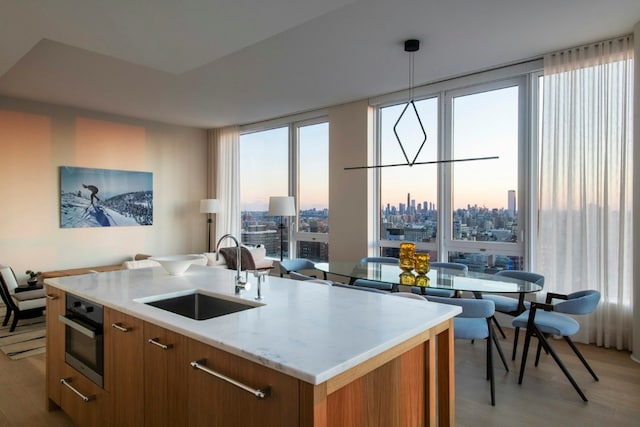 kitchen featuring an island with sink, stainless steel oven, decorative light fixtures, sink, and a healthy amount of sunlight