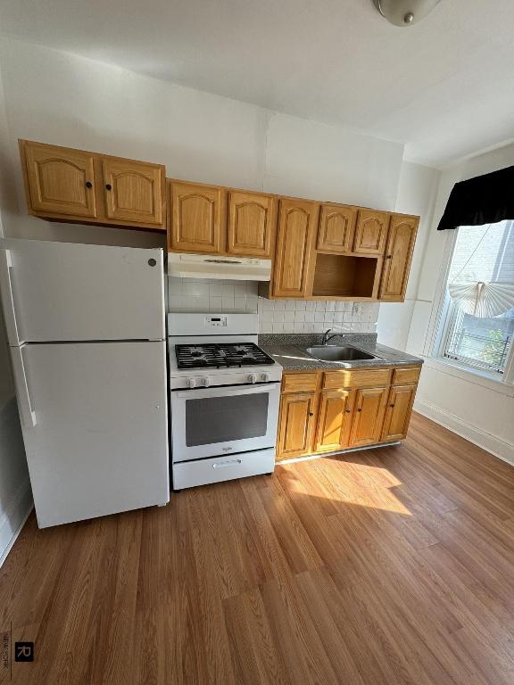 kitchen featuring under cabinet range hood, white appliances, wood finished floors, a sink, and dark countertops