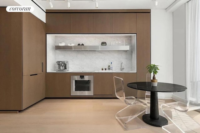 bar featuring rail lighting, tasteful backsplash, ventilation hood, stainless steel oven, and light wood-type flooring