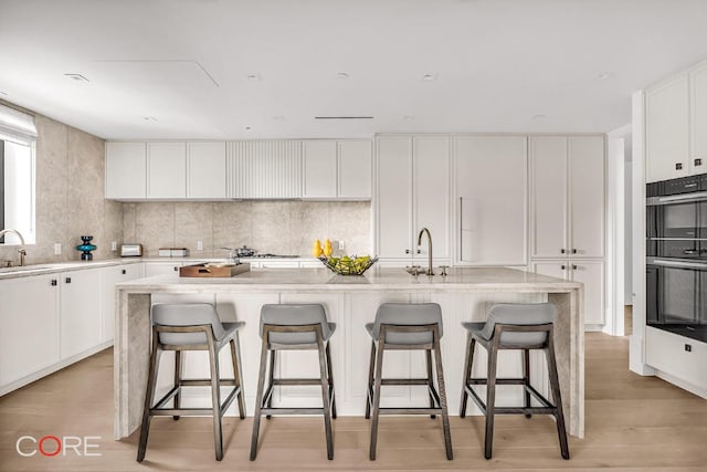 kitchen featuring white cabinetry, multiple ovens, a kitchen bar, and a kitchen island with sink