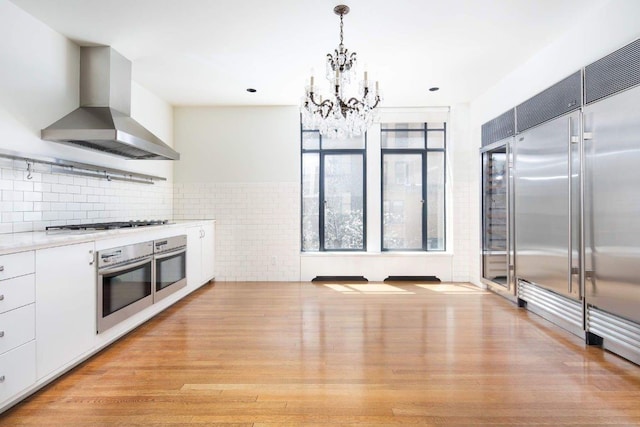 kitchen with white cabinetry, decorative light fixtures, light wood-type flooring, appliances with stainless steel finishes, and range hood