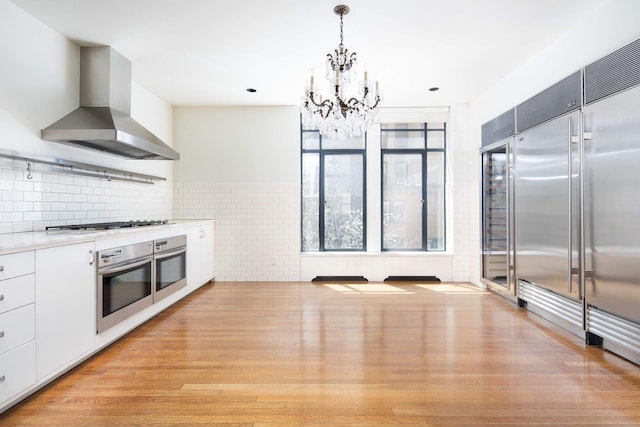 47 E 91st St Unit 2, New York City, NY 10128 - kitchen featuring a notable chandelier, light wood finished floors, white cabinetry, appliances with stainless steel finishes, and wall chimney range hood