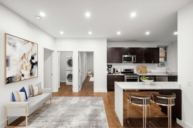 kitchen with stacked washer / drying machine, light hardwood / wood-style floors, dark brown cabinetry, appliances with stainless steel finishes, and a breakfast bar area