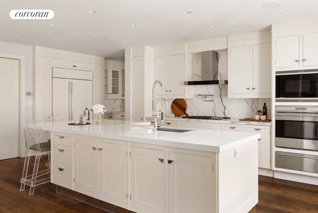 kitchen featuring a warming drawer, light countertops, a kitchen island with sink, wall chimney range hood, and built in appliances