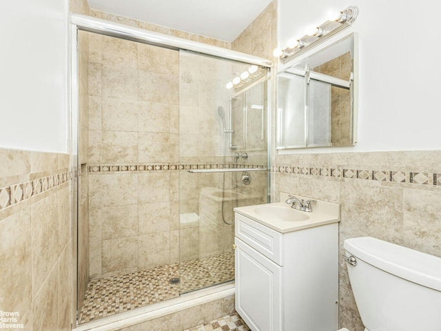 bathroom with a stall shower, toilet, a wainscoted wall, vanity, and tile walls