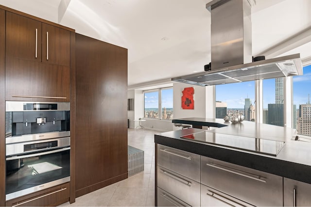 kitchen with dark brown cabinets, black electric stovetop, island range hood, light tile patterned flooring, and stainless steel double oven