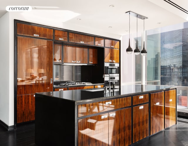 kitchen with an island with sink, sink, dark stone counters, hanging light fixtures, and stainless steel appliances