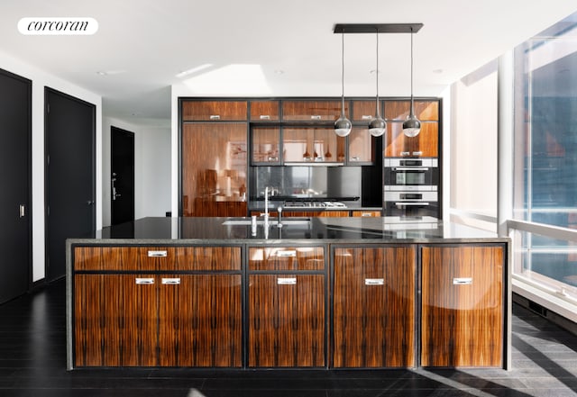 kitchen featuring sink, dark stone countertops, double oven, a center island with sink, and decorative light fixtures