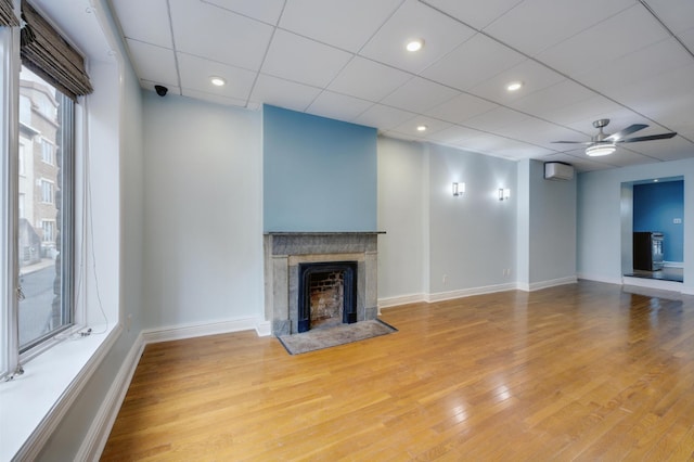 unfurnished living room with a drop ceiling, a wall unit AC, ceiling fan, and light wood-type flooring