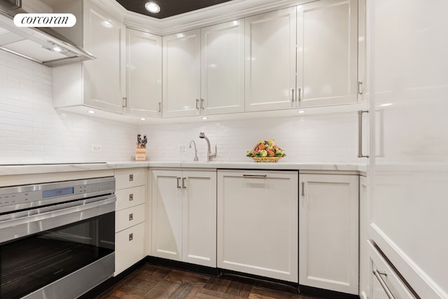 kitchen with dark parquet flooring, sink, white cabinetry, stainless steel oven, and wall chimney range hood