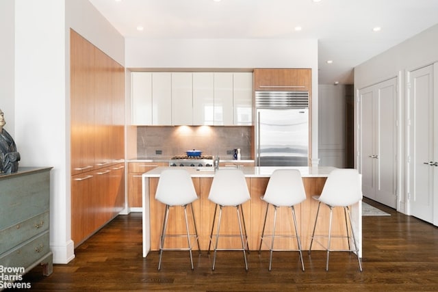 kitchen with a kitchen island with sink, a breakfast bar, white cabinetry, light countertops, and stainless steel built in refrigerator