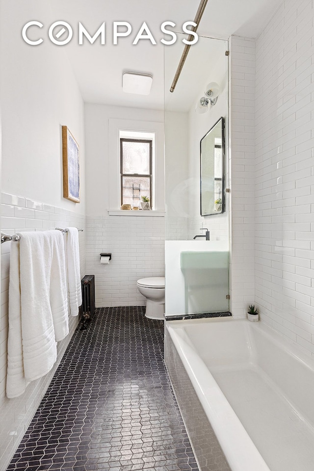 bathroom with a tub to relax in, a wainscoted wall, tile walls, toilet, and tile patterned floors