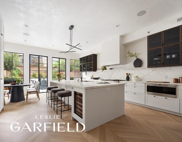 kitchen with beverage cooler, decorative backsplash, hanging light fixtures, light parquet flooring, and a center island with sink