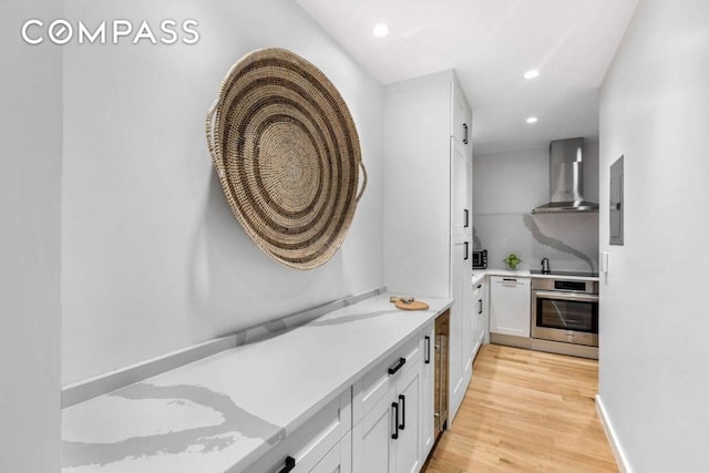 kitchen featuring light wood-style floors, white cabinetry, stainless steel oven, wall chimney range hood, and dishwasher