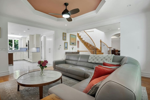 living room featuring light wood finished floors, stairway, a tray ceiling, ornamental molding, and arched walkways