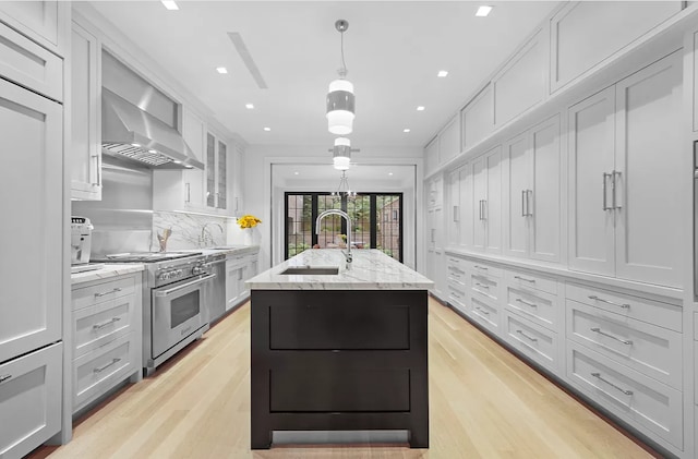 kitchen with light stone counters, a sink, wall chimney range hood, stainless steel range, and a center island with sink