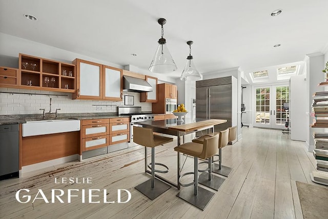 kitchen featuring wall chimney range hood, backsplash, high quality appliances, decorative light fixtures, and light wood-type flooring