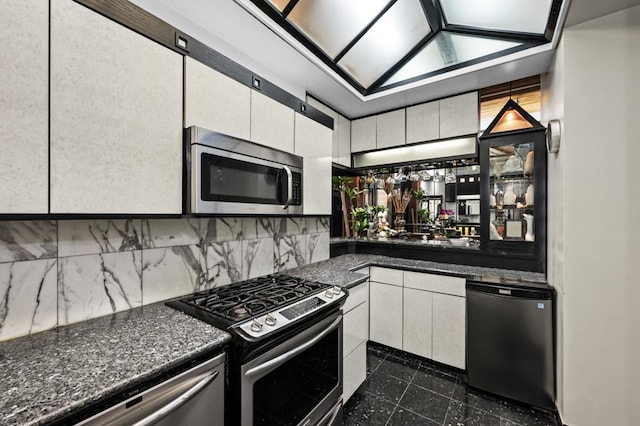 kitchen with stainless steel appliances, dark stone countertops, backsplash, and white cabinetry