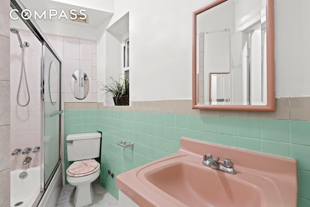 bathroom featuring a wainscoted wall, tile walls, toilet, enclosed tub / shower combo, and a sink