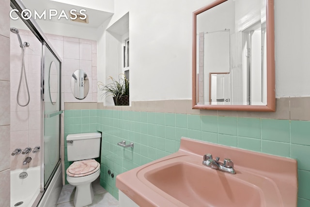 bathroom with tile walls, a sink, a wainscoted wall, toilet, and enclosed tub / shower combo