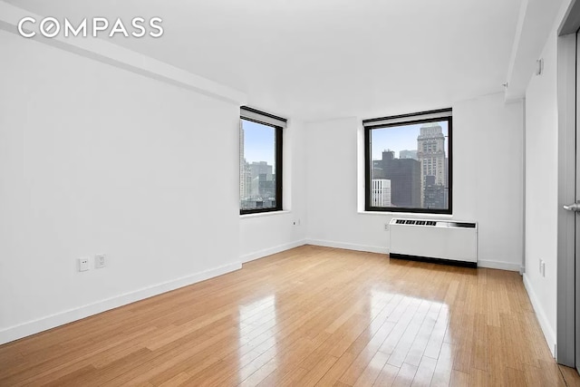 unfurnished bedroom with light wood-type flooring, a view of city, radiator, and baseboards