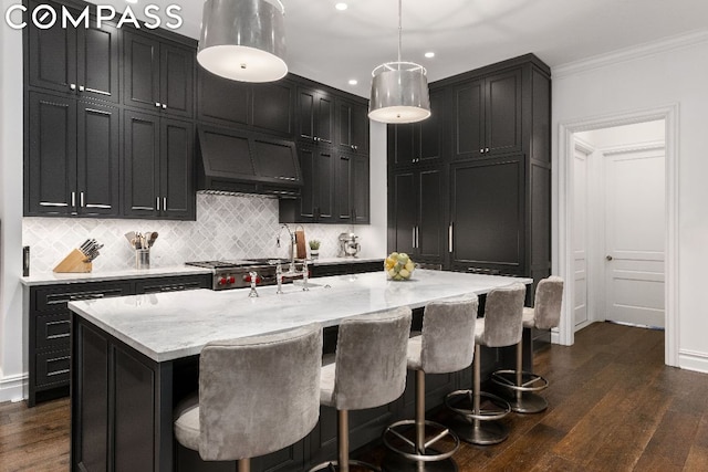 kitchen with dark hardwood / wood-style flooring, premium range hood, hanging light fixtures, a kitchen island with sink, and crown molding