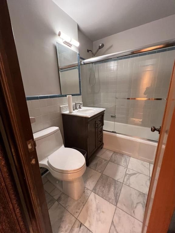 full bathroom featuring toilet, bath / shower combo with glass door, tile walls, and vanity