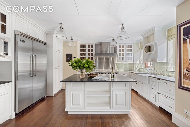 kitchen with dark countertops, wall chimney exhaust hood, white cabinetry, and built in appliances