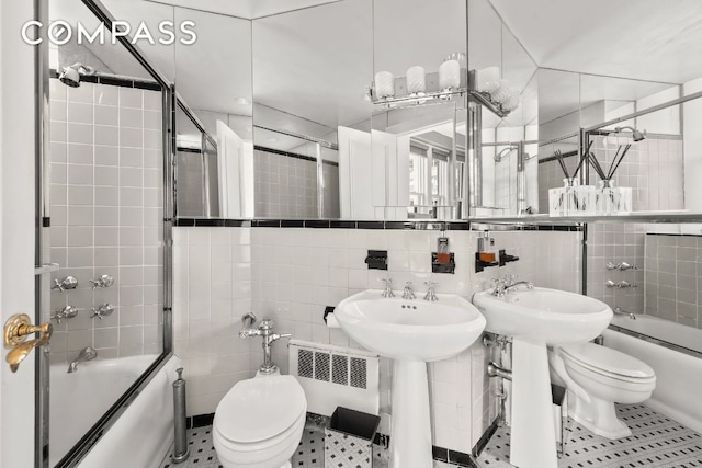 bathroom featuring toilet, radiator heating unit, bath / shower combo with glass door, and tile walls