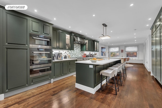 kitchen featuring an island with sink, light countertops, green cabinets, a warming drawer, and wall chimney exhaust hood